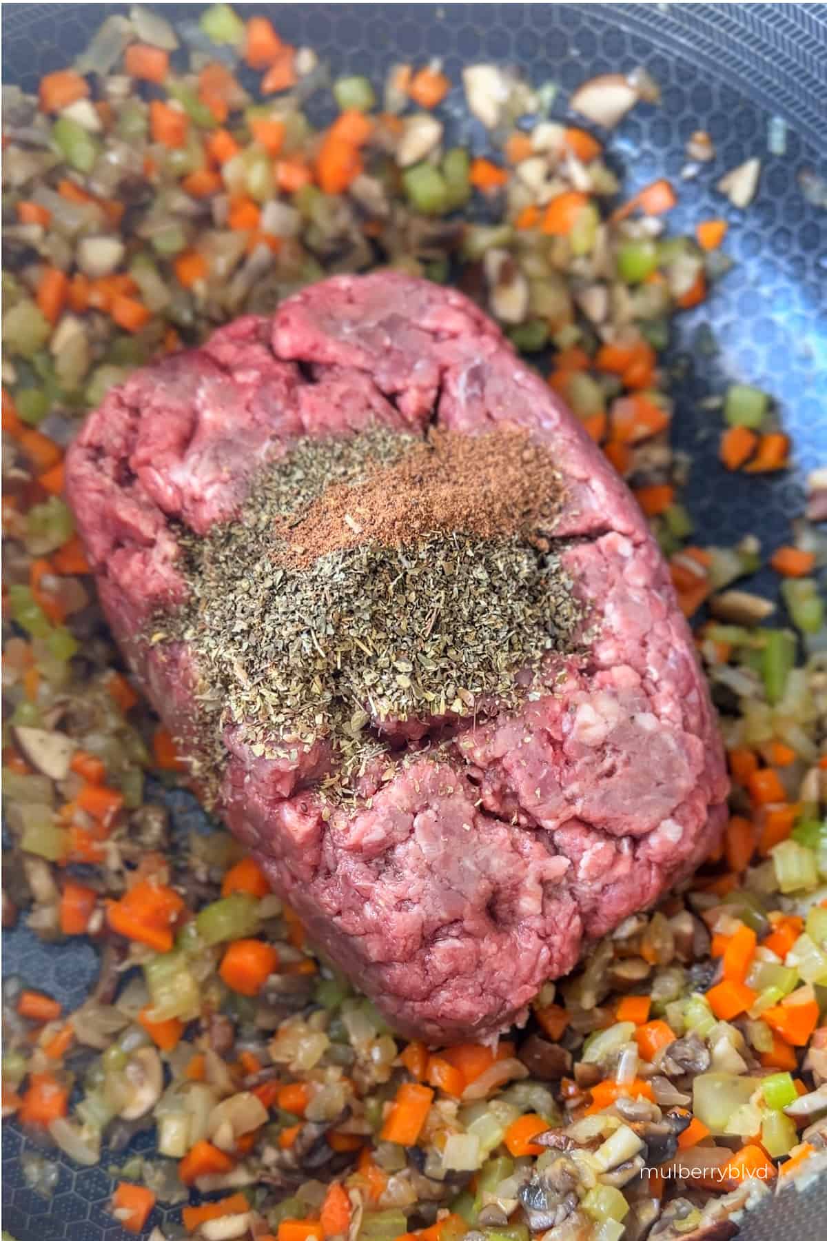an image of raw ground beef with nutmeg, oregano, and basil on top, being added to sautéed finely chopped mushrooms, onions, celery and carrots