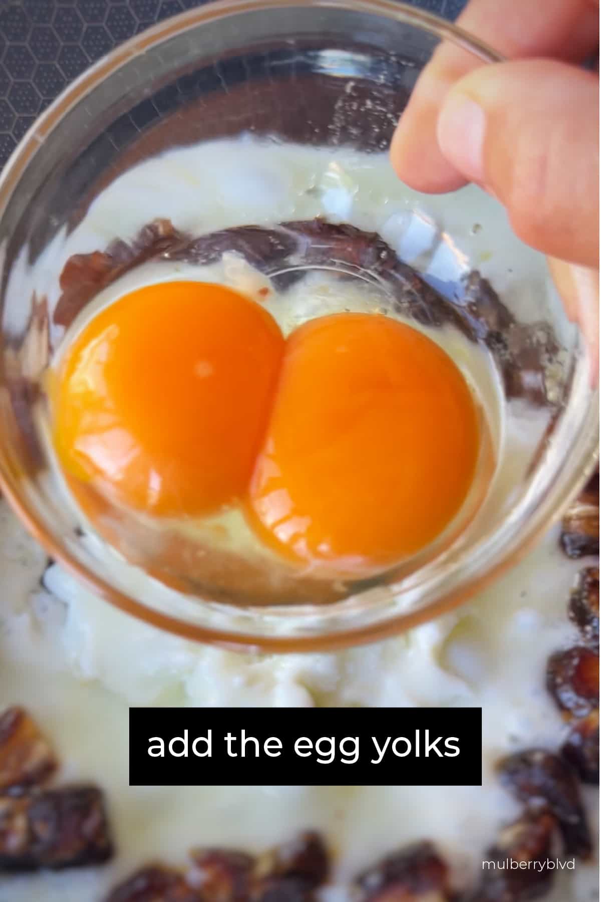 This image shows a hand holding a a small bowl with two egg yolks, with the image saying "add the egg yolks".
