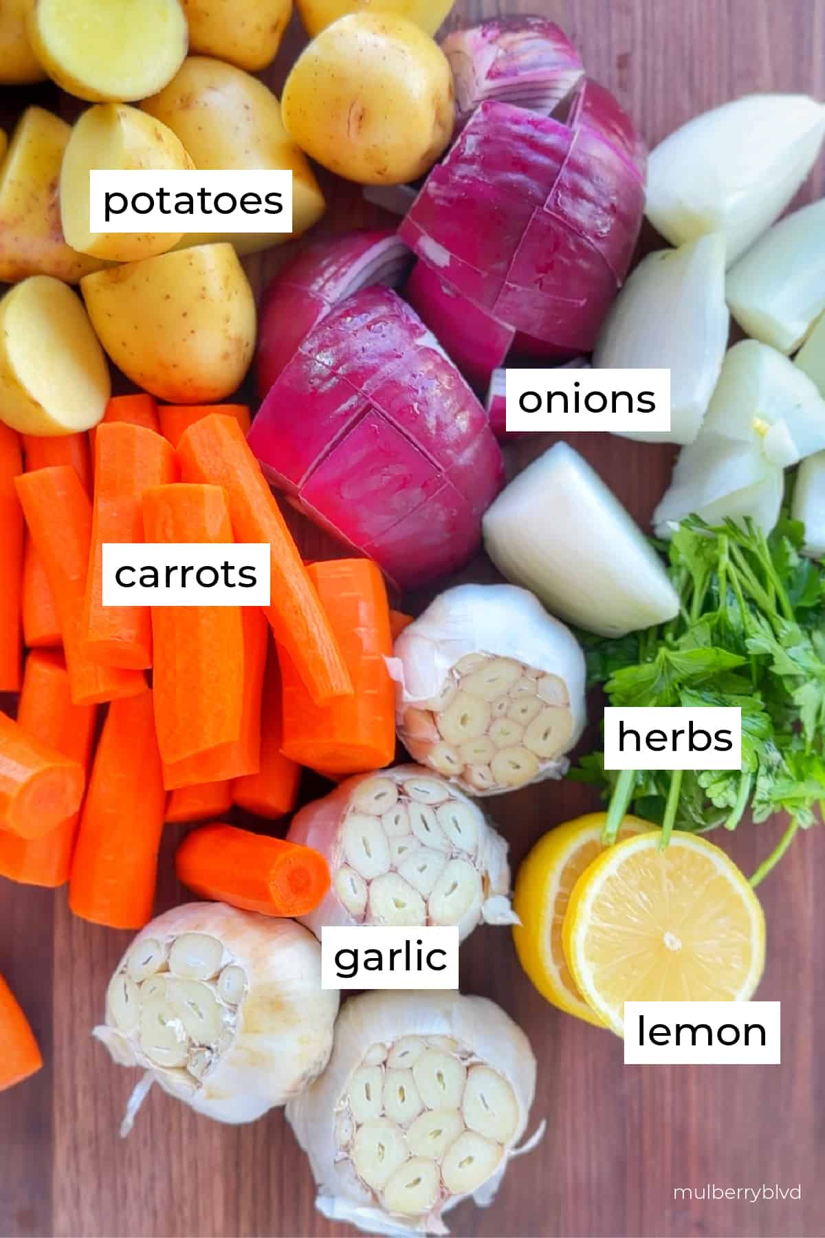 This is an image of cut and prepped garlic, lemon, herbs, carrots, potatoes, and both red and white onions.