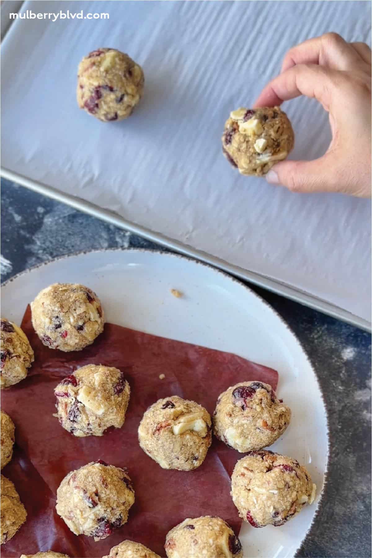 Oatmeal cookie dough with white chocolate chips, dried cranberries, and toasted pecans, being placed on a cookie sheet.