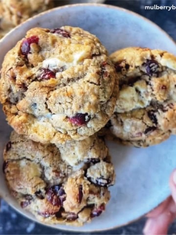 Oatmeal cookies with white chocolate chips, dried cranberries, and toasted pecans, stacked on a plate and perfect for holiday baking.